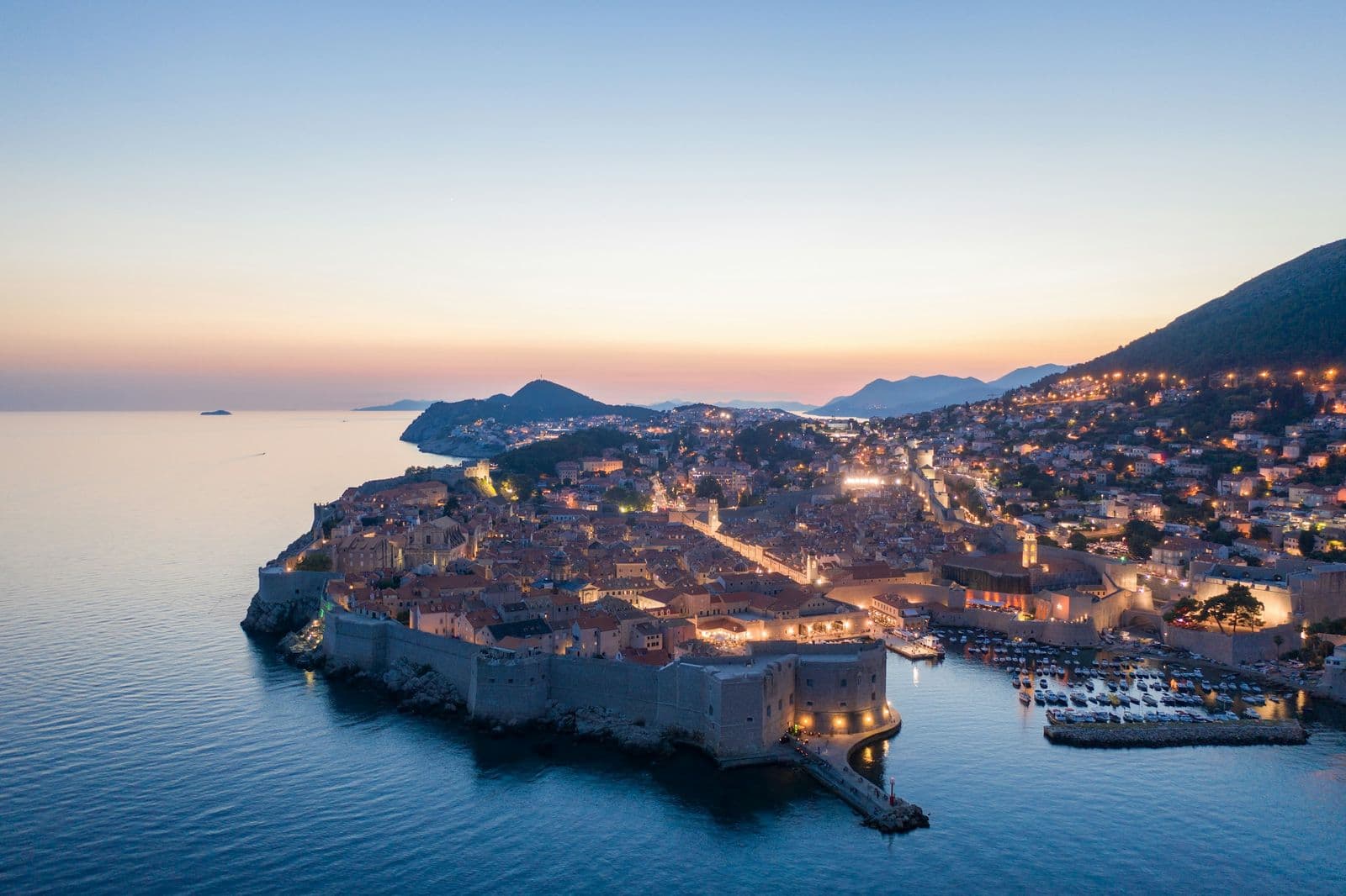 The walled Old Town of Dubrovnik from the air at dusk, its walls and harbour lit